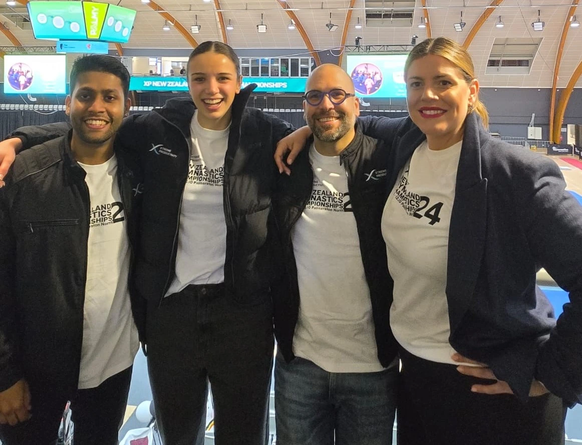 Four people standing at the gymastics championships. they are all smiling and have their arms around each other. Wearing white XP Gymnastics championships tees